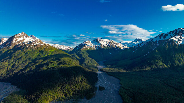 Aerial view of mountain valley and river in Alaska. Alaskan river flowing through deep green valley and snow-capped mountains under blue sky.