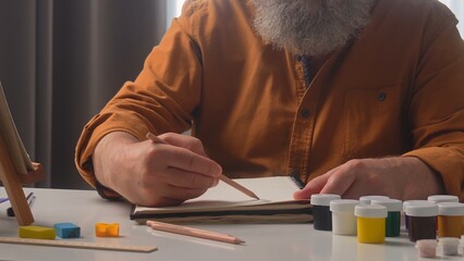 An elder male artist sketches carefully on a notepad at home, surrounded by art supplies and paint...