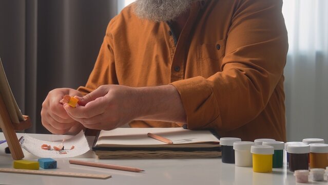 A senior male artist is depicted at home, deeply focused on his creative work with art materials.