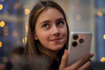 Young smiling happy woman wear casual clothes hold in hand use mobile cell phone. A teenage girl with a smartphone in her hand on a dark blurred background with Christmas lights