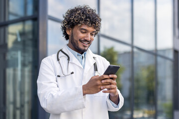 Close-up photo of a young Indian male doctor in a white coat standing near a hospital and using a mobile phone