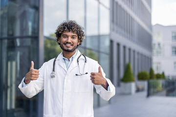Portrait of a smiling young Indian male doctor, student standing outside the clinic in a white coat and showing the super sign with his fingers at the camera