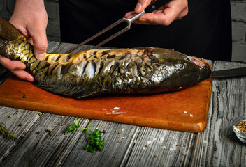 A person skillfully holds a large fish on a wooden cutting board, preparing it for cooking. Fresh herbs and seasoning are scattered around in a rustic kitchen © Віталій Б.