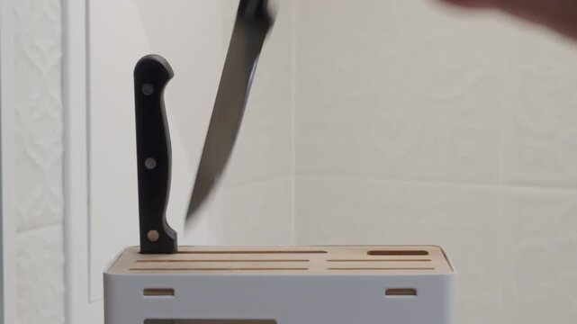 A knife block, featuring a light wood interior and a white exterior, holds a set of knives with black handles and a sharpening tool, standing upright against a light-colored, textured kitchen wall