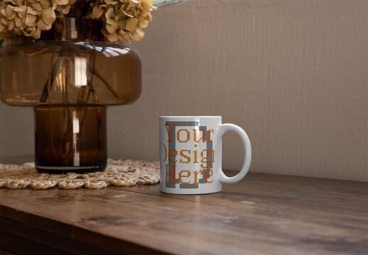 Mockup of white cup sits on a wooden table next to a large glass vase with dried flowers.