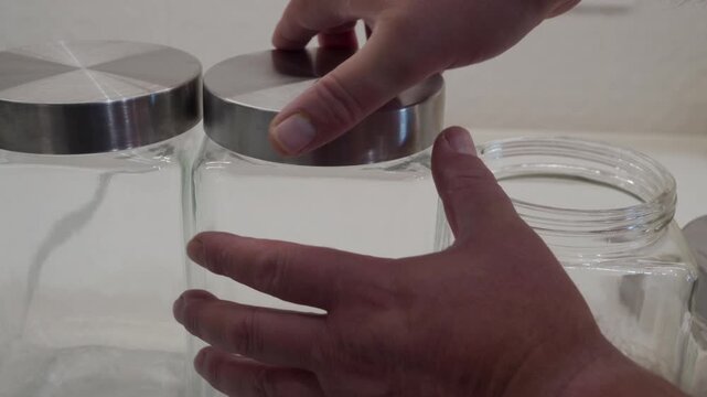 A hand adjusts a metal lid on a clear glass storage jar, sitting with another on a white countertop. This highlights an act of meticulous kitchen organization and preparation.