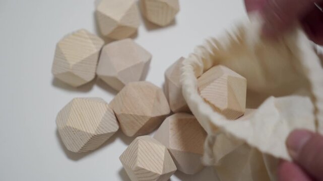 Natural wood faceted building blocks are scattered on a white surface, with a hand holding a drawstring bag nearby. This captures a moment of play or tidying up.
