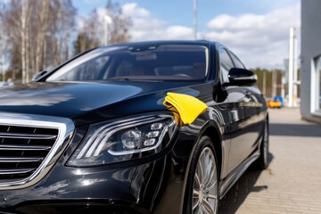 A sleek black luxury car being polished with a yellow cloth in a sunny outdoor setting