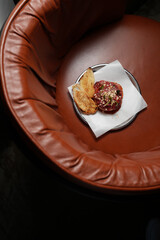 An overhead, vertical shot of a gourmet beef tartare served on a small metal plate with toasted crostini slices, resting on the rich brown leather seat of a stylish armchair