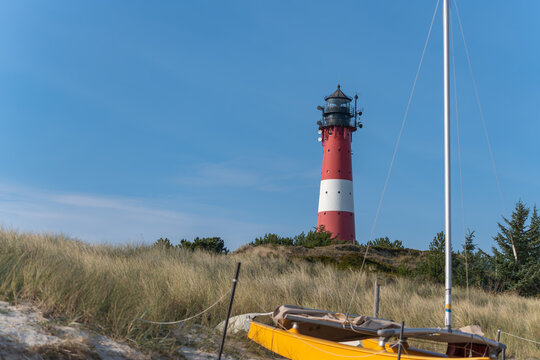 A yellow boat rests in the sand dunes with the lighthouse in the distance. A calm coastal scene under a blue sky conveys tranquility. - Powered by Adobe