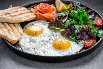Fried eggs with salmon and salad served with toast. Breakfast with fried eggs, salmon, red caviar, salad, and toasted bread on a black plate.