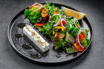 Greek salad with feta and olives on black plate. Fresh Greek salad with feta cheese, olives, bell peppers, tomatoes, and lemon served on a black plate.