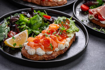 Bruschetta with salmon, avocado, and salad greens. Close-up of salmon bruschetta with avocado slices, red caviar, and salad greens on a black plate.