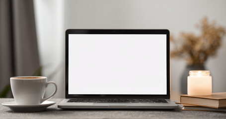 Mockup of a laptop with a blank screen on a cozy home office desk. The setup includes a white coffee cup and a lit candle on books, conveying a comfortable, relaxed, and focused working environment