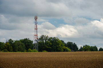 Obraz premium Telecommunication tower standing tall in an open agricultural field against a backdrop of green trees and white clouds. A symbol of modern technology integrated into a rural landscape.