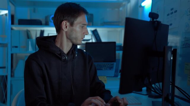 Professional male programmer working night shift in IT room, coding and testing software on multiple monitors, analyzing project data and debugging code, showcasing innovation and technology.