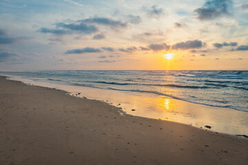 Sunset over stormy North Sea beach with reed grass bushes, orange-red sky over the dark blue ocean, panorama with sandy beach and blowing grass in the wind, storm at sunset over wavy water
