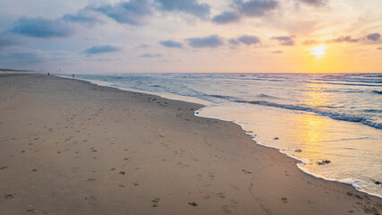 Sunset over stormy North Sea beach with reed grass bushes, orange-red sky over the dark blue ocean, panorama with sandy beach and blowing grass in the wind, storm at sunset over wavy water