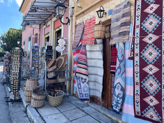 Colorful Greek street souvenir shop with handmade woven rugs, baskets, and traditional crafts hanging outside. Bright textiles, pink building facade, and sunny Mediterranean atmosphere.