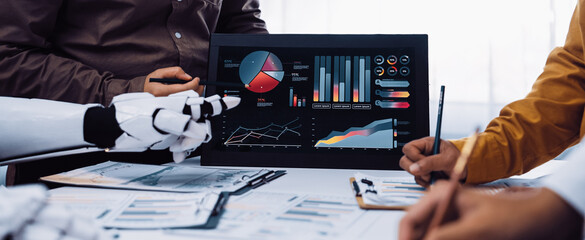 A diverse team collaborates in a modern office, discussing data analysis and insights. The robot interacts with digital displays featuring charts and graphs.Noogenesis