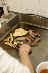 A person is carefully grilling a variety of cuts of meat on a flat cooking surface located in a kitchen