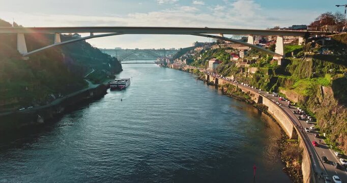 Portugal, Porto: Douro River with cruise boats, bridges and car traffic moving along the riverbank, showcasing urban city landscape with green hills under clear sky. Aerial view drone footage