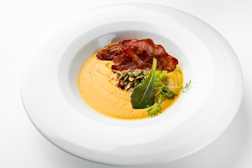 Gourmet pumpkin soup with crispy bacon, seeds, and fresh herbs served in a white bowl. Autumn-themed dish with elegant plating, ideal for restaurant or seasonal food concepts.