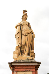 female beige statue with cape and helmet on a red stone base on a white background