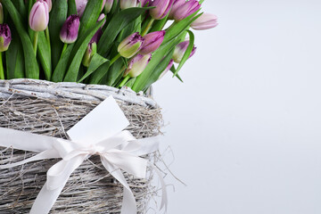 Purple and pink tulips in rustic white wicker basket with white ribbon and blank gift tag on light background