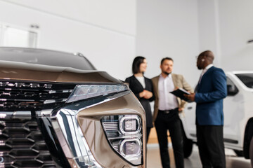 Close-up of modern vehicle front with sleek design, while three professionals engage in discussion about automotive features and sales strategies in a bright showroom environment