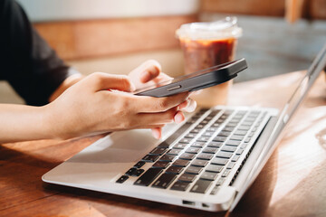 Remote work concept. Hands Holding Smartphone While Working on Laptop in Cozy Coffee Shop Atmosphere with Cold Brew Drink Nearby