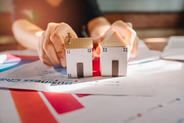 Real Estate concept. Hands Comparing Two Miniature Houses on Business Charts with Colorful Graphs and Diagrams in Bright Office Setting