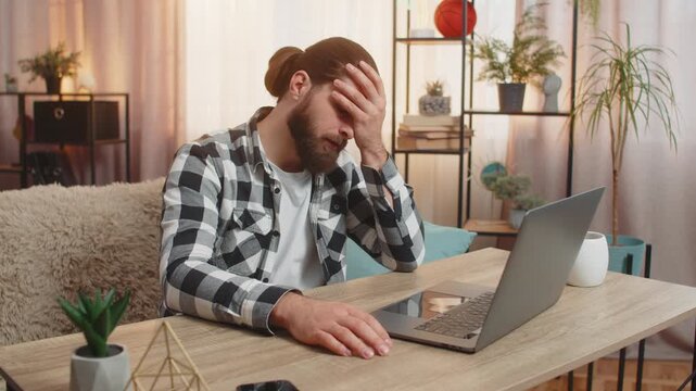 Man concentrates at home table, typing on laptop until stumbling upon a big glitch error virus that sparks immediate annoyance. Guy slaps forehead, quietly grumbling at unexpected technical issue fail