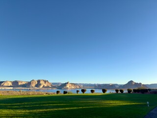Obraz premium Serene landscape view of Lake Powell featuring vast water expanse against a backdrop of majestic sandstone rock formations under clear blue sky and lush green foreground