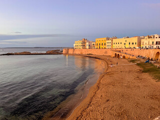 Abendstimmung an der Stadtmauer von Gallipoli in Apulien, Italien. Goldene Sonne beleuchtet die historischen H&auml;user am Sandstrand mit ruhigem Meer.