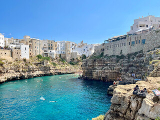 K&uuml;stenstadt Polignano a Mare in Apulien, Italien &ndash; wei&szlig;e H&auml;user auf steilen Klippen &uuml;ber t&uuml;rkisblauem Meer, Badeg&auml;ste im klaren Wasser, Sommerstimmung unter blauem Himmel.