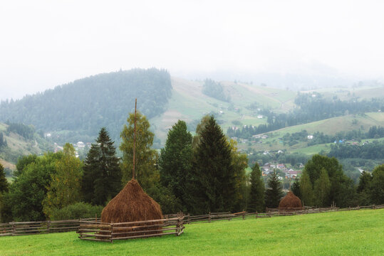 Idylic foggy rural mountain landscape in Carpathians. Traditional haystacks standing on a green pasture overlooking misty  hills  covered with forest and rural settlements in the distant background.