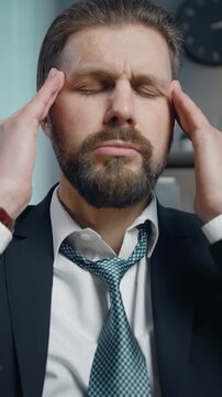 A tired businessman sits in his office, visibly stressed. He closes his eyes and massages his forehead, seeking relief from his busy workday and finding a moment of peace.