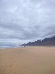 Beautiful Cofete Beach on Fuerteventura, Canary Islands. Wide sandy coastline with dramatic cloudy mountains in the background and the Atlantic Ocean waves in soft light.