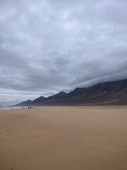 Beautiful Cofete Beach on Fuerteventura, Canary Islands. Wide sandy coastline with dramatic cloudy mountains in the background and the Atlantic Ocean waves in soft light.