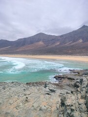 Beautiful Cofete Beach on Fuerteventura, Canary Islands. Wide sandy coastline with dramatic cloudy mountains in the background and the Atlantic Ocean waves in soft light.