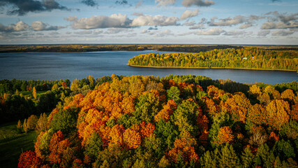 Aerial view of a lake surrounded by a colorful autumn forest, with golden, orange, and green trees...