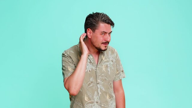 Middle-aged man trying hear you, looking confused frowning, keeping arm near ear for louder voice, asking to repeat, to hear information, deafness. Young adult guy shows confusion on blue background.