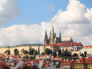 Obraz premium Majestic gothic cathedral rising above historic red rooftops under a bright blue sky with fluffy white clouds during a sunny autumn afternoon showing intricate stone architecture and towers.