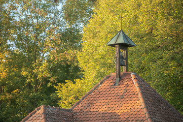 Glockenstuhl mit grünem Kupferdach und Kreuz auf Walmdach mit roten Dachziegeln vor Bäumen mit Herbstlaub in Reutlingen-Mittelstadt im Streiflicht