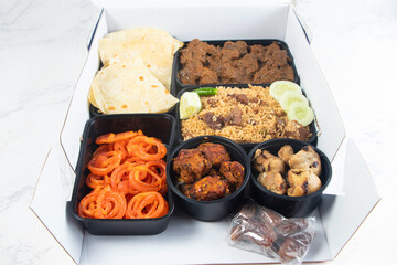 Iftar Meal Box South Asian Catering with Paratha Slice, Meat Biryani, Beef Curry, Flatbread, Chicken Malai Tikka Boti, and Jalebi isolated on marble background top view closeup