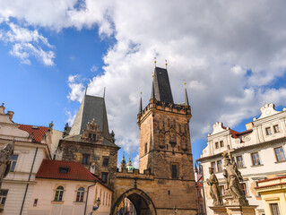 Fototapeta premium Majestic gothic stone tower and historical bridge entrance with classic statues and elegant white buildings under a bright blue sky with white clouds in an old town square.