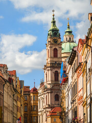 Obraz premium Vertical view of an ornate baroque church tower with a green copper dome and a large black clock face standing against a blue sky with soft white clouds and sunlit facades.