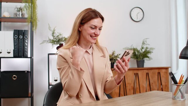 Middle-aged businesswoman at home office wins discount in app on smartphone victory, positive good news of salary raise. Freelancer girl smiles at table celebrates win checks code payment success big
