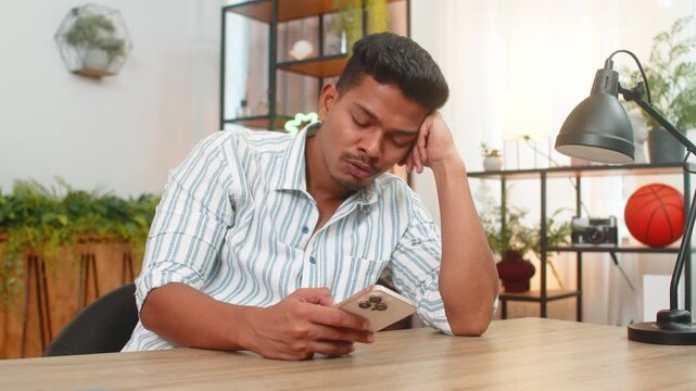 Indian man at home scrolls smartphone news feels tired bored drained by long feed with ads. Arabian guy at table looks away breathes slow mutes alerts puts phone aside and plans short break for calm.
